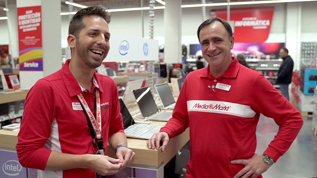 Dos hombres con camisas rojas están sonriendo en una tienda. Hay computadoras portátiles en exhibición. Se ven carteles en rojo y blanco.