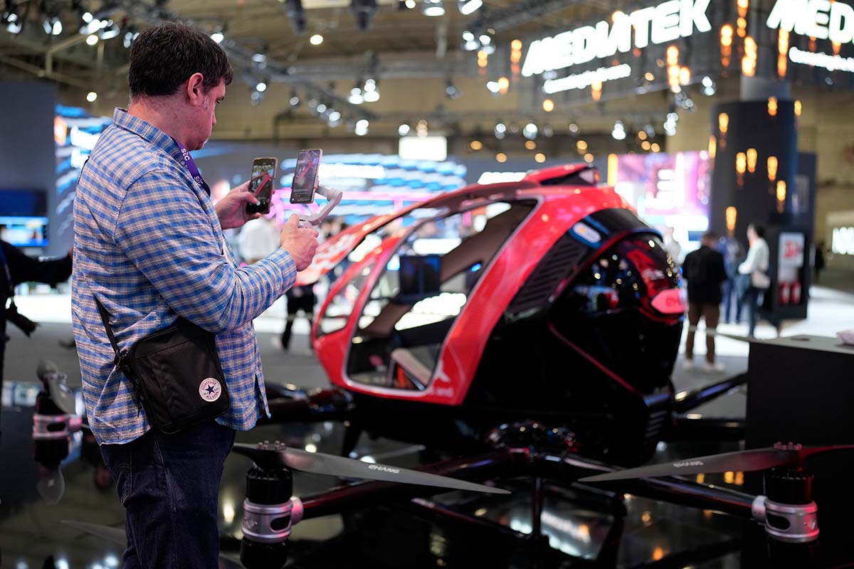 Un hombre usa dos teléfonos frente a un dron rojo y negro en una exposición. El fondo muestra letreros de "MEDIATEK" y más personas.