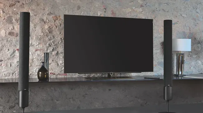 En un entorno moderno, un televisor de pantalla plana se sitúa entre dos altavoces cilíndricos negros. A la derecha, una lámpara de mesa blanca.