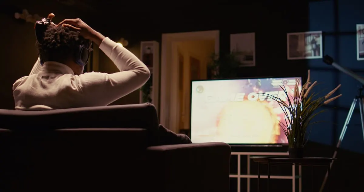 Persona sentada de espaldas en un sillón, con auriculares y las manos en la cabeza, mirando una pantalla de televisor con el mensaje “Game Over”.