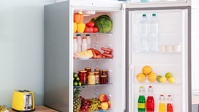 Un refrigerador blanco abierto muestra verduras, frutas, leche, queso, embutidos y tarros en los estantes. Una tostadora amarilla está cerca.
