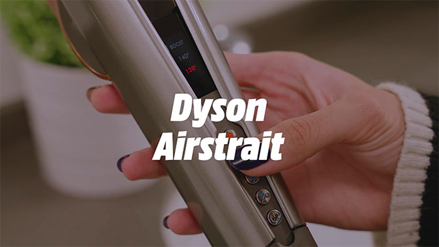 Mano sujetando una plancha Dyson Airstrait gris. La plancha tiene una pantalla y varios botones. Al fondo, una planta en una maceta blanca.