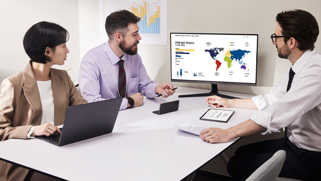 Tres personas en una mesa blanca con un monitor mostrando un mapa mundial y gráficos de barras, además de una laptop.
