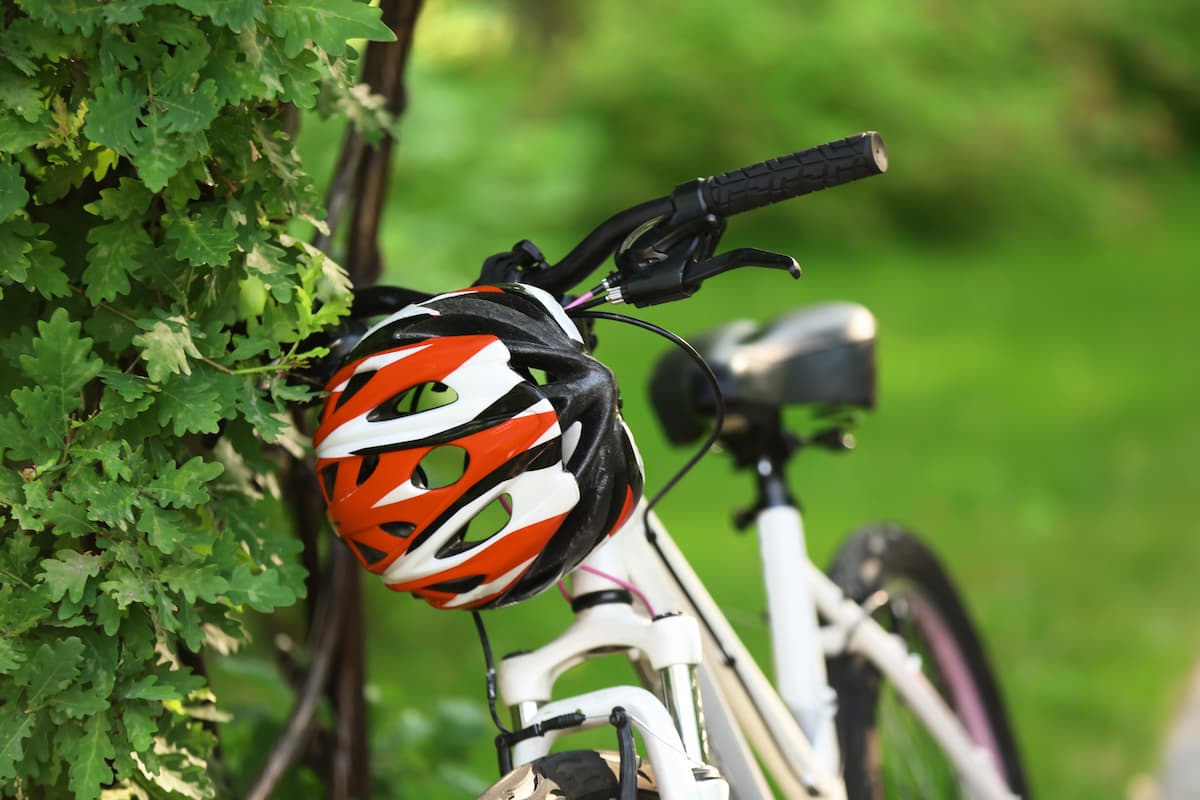 Casco de ciclismo naranja y blanco colgado en el manillar de una bicicleta junto a vegetación verde.