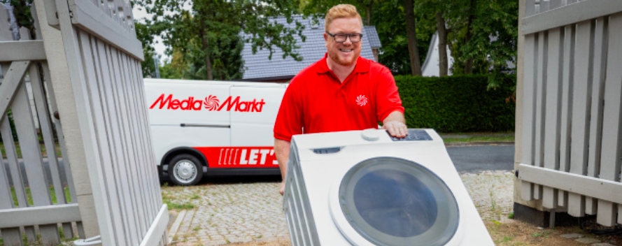  Empleado de MediaMarkt entrega una lavadora a domicilio, empujándola por el jardín con una furgoneta de la marca al fondo.