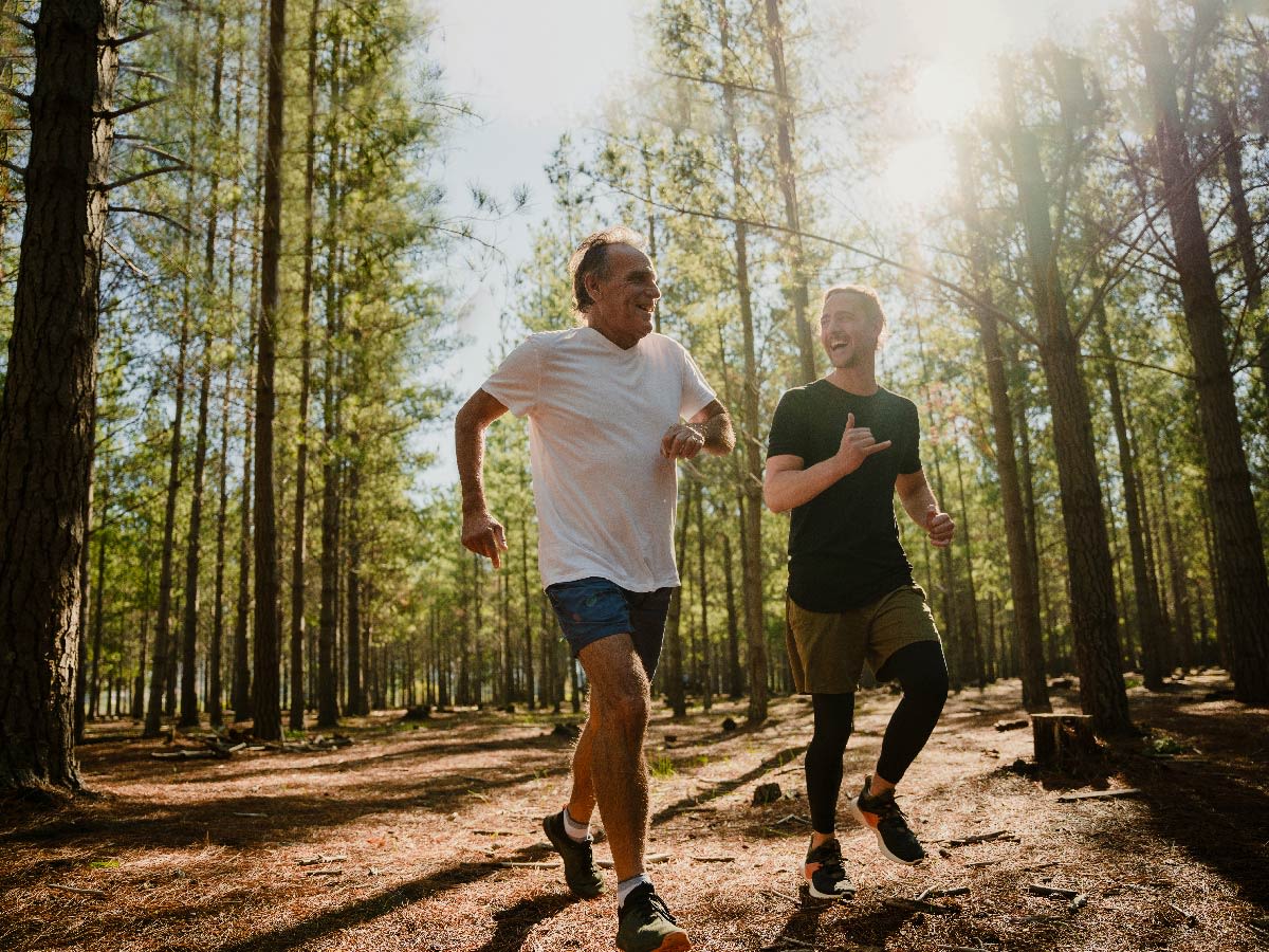 Dos personas corriendo por un sendero en un bosque soleado.

