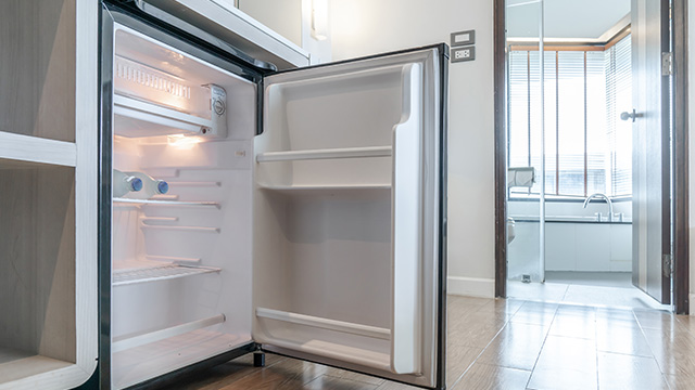 Un refrigerador blanco con la puerta abierta y estantes vacíos. Al fondo, un baño con puerta de cristal.