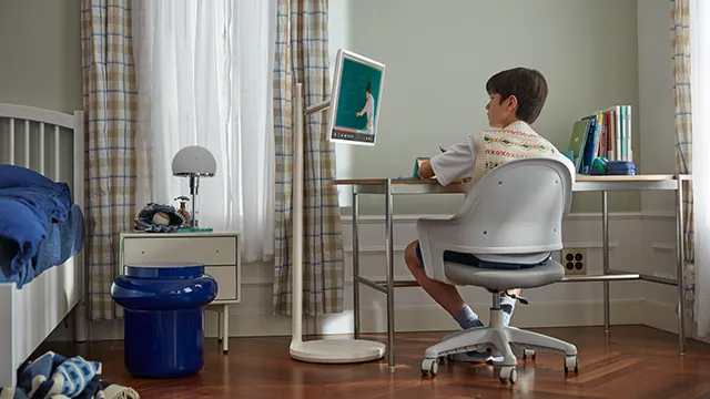 Un niño está sentado en un escritorio gris frente a una pantalla. Hay cortinas y una cama en el fondo.