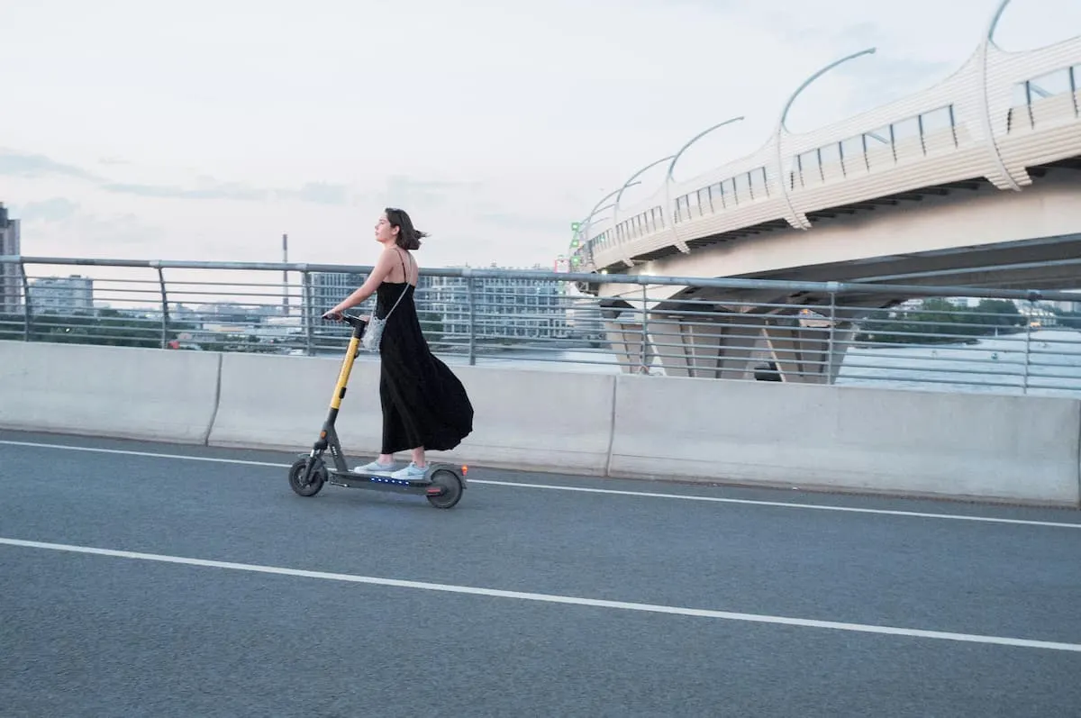 Imagen de una chica morena con el pelo corto y vestido largo negro yendo en patinete eléctrico por una carretera. Fondo de un puente con vistas a una ciudad.