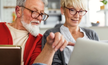 Una pareja mayor con gafas mira un portátil. El hombre, con barba blanca, viste una camisa roja. Hay un mensaje de verificación verde.