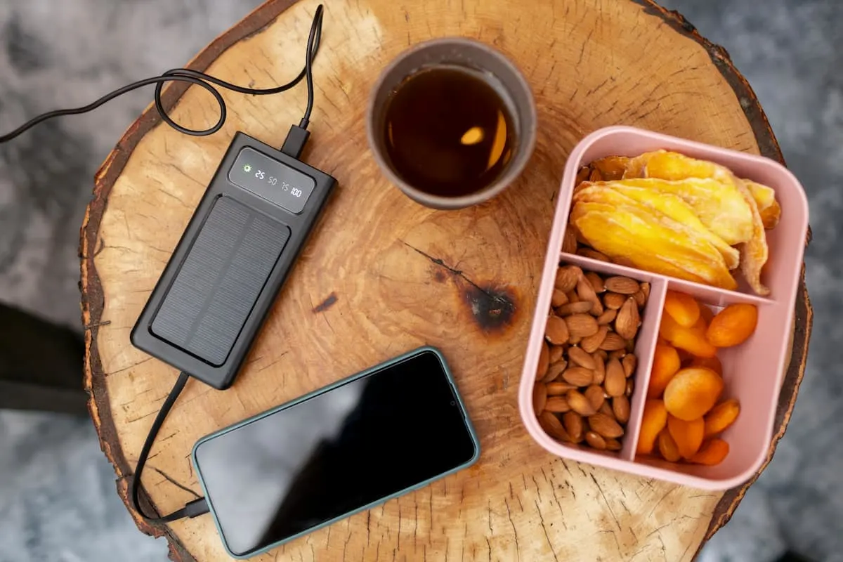 Mesa de madera con un móvil conectado a un powerbank solar, un vaso con bebida y un recipiente con frutos secos y fruta deshidratada.