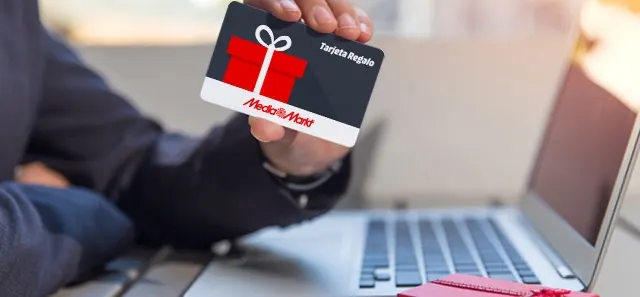 Una mano sostiene una tarjeta regalo gris, blanca y roja de Media Markt frente a un portátil abierto.