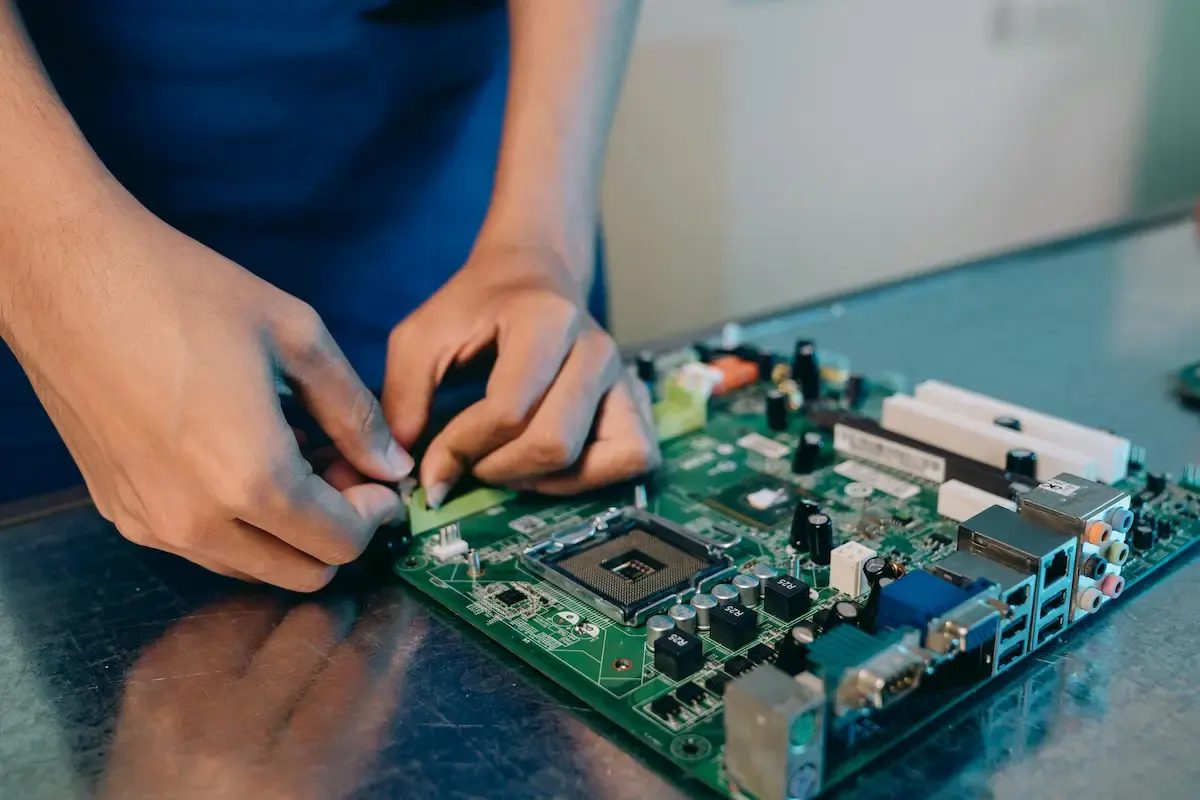 Manos conectando un componente a una placa base verde sobre una mesa metálica, con fondo blanco.