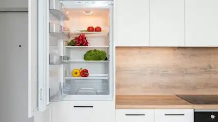 Vista de una cocina moderna con nevera abierta. En los estantes hay tomates, rábanos, lechuga y pimientos. Los muebles son blancos y de madera.