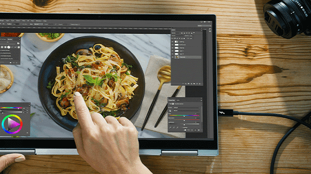 Ordenador portátil con plato de pasta en pantalla, mano tocando, sobre mesa de madera con cámara y cable.