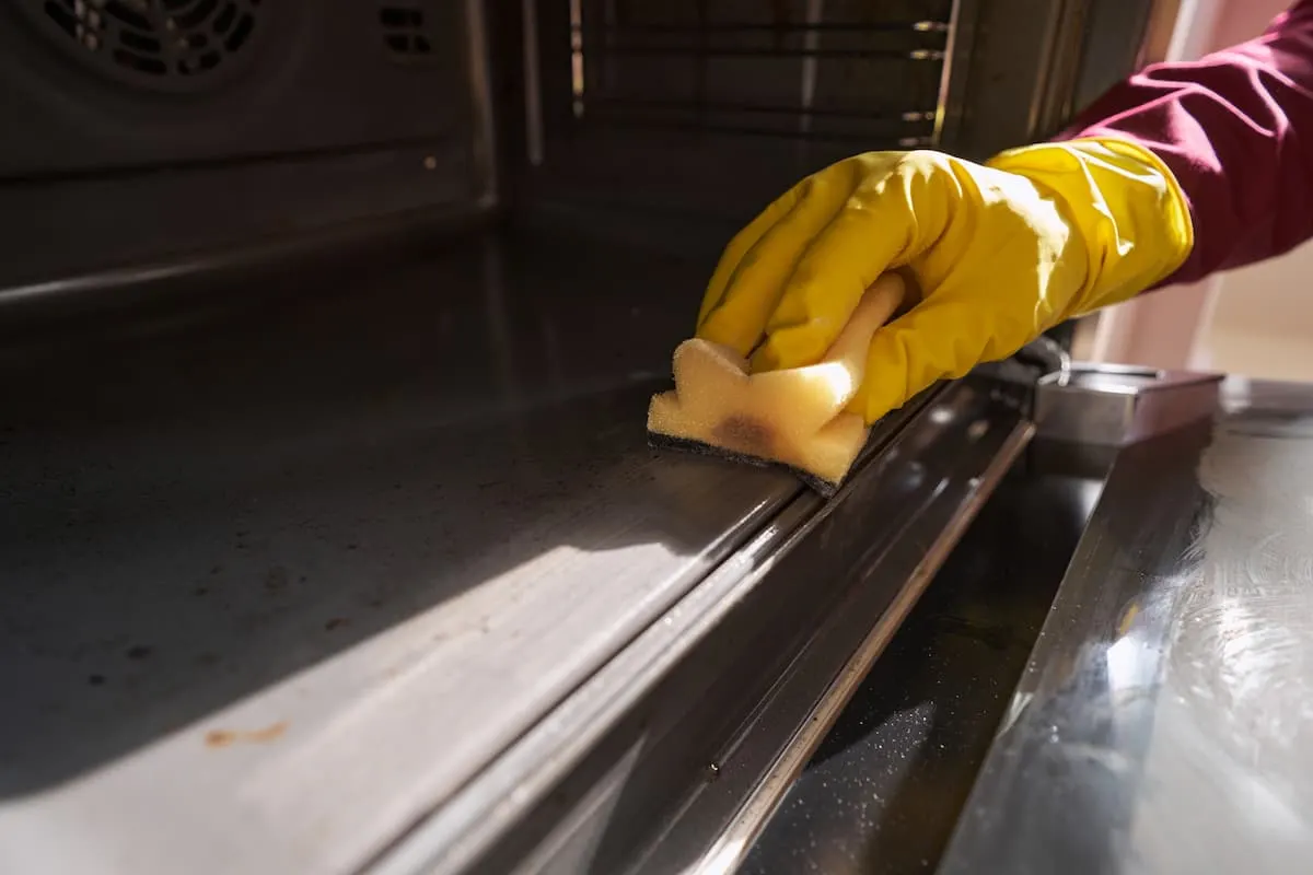 Mano con guante amarillo de goma limpiando con una esponja el riel interior de un horno de acero inoxidable.