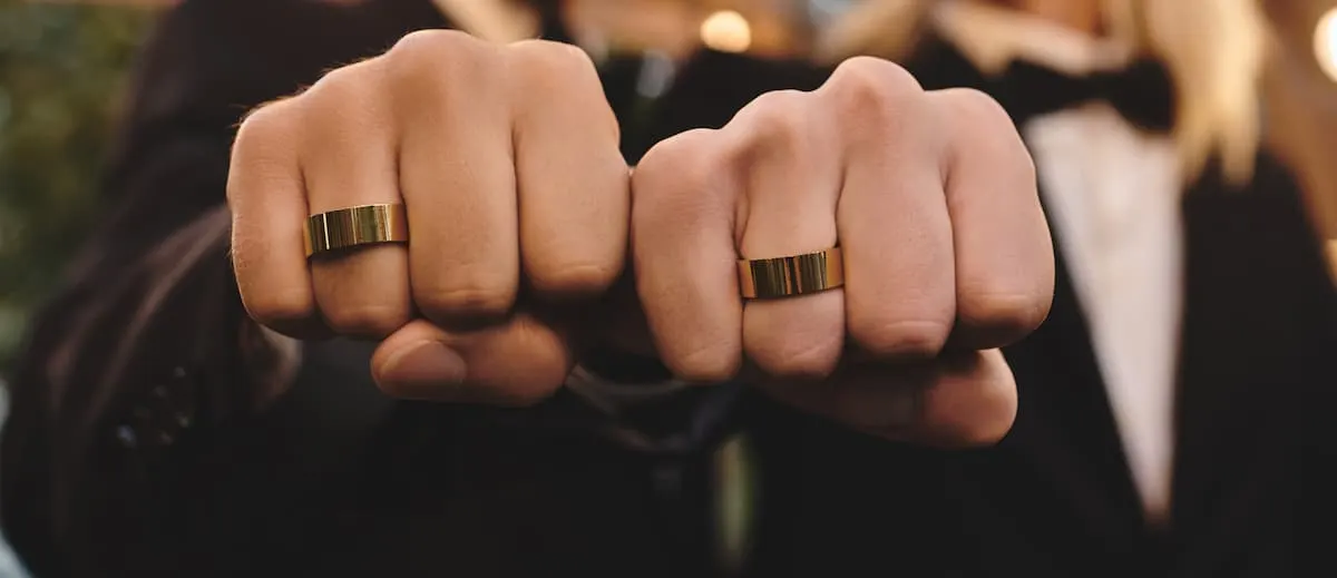 Dos personas con trajes elegantes muestran sus manos cerradas, luciendo anillos dorados idénticos en los dedos corazón.