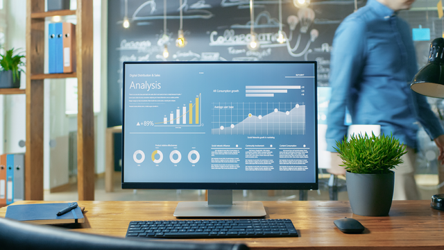 Escritorio con pantalla que muestra gráficos de análisis, teclado, planta y libreta. Persona borrosa al fondo.