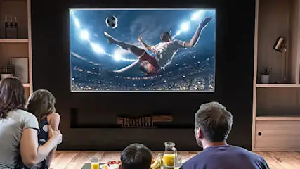 Familia mirando un partido de fútbol en la televisión en una sala de estar con estanterías y suelo de madera.