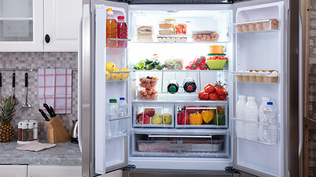 Frigorífico abierto lleno de alimentos como frutas, verduras, huevos y bebidas, en un ambiente de cocina con armarios blancos.