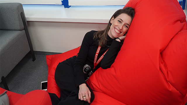 Mujer sonriente descansa en un puff rojo. Lleva ropa negra y un cordón rojo. Un sofá gris está al fondo.