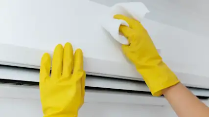 Manos enguantadas en amarillo limpian una superficie blanca con un paño blanco. Una mano sostiene la superficie, la otra limpia.