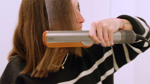 Mujer usa plancha de pelo naranja y gris sobre un mechón. Lleva un suéter a rayas blancas y negras y un collar. Fondo blanco.