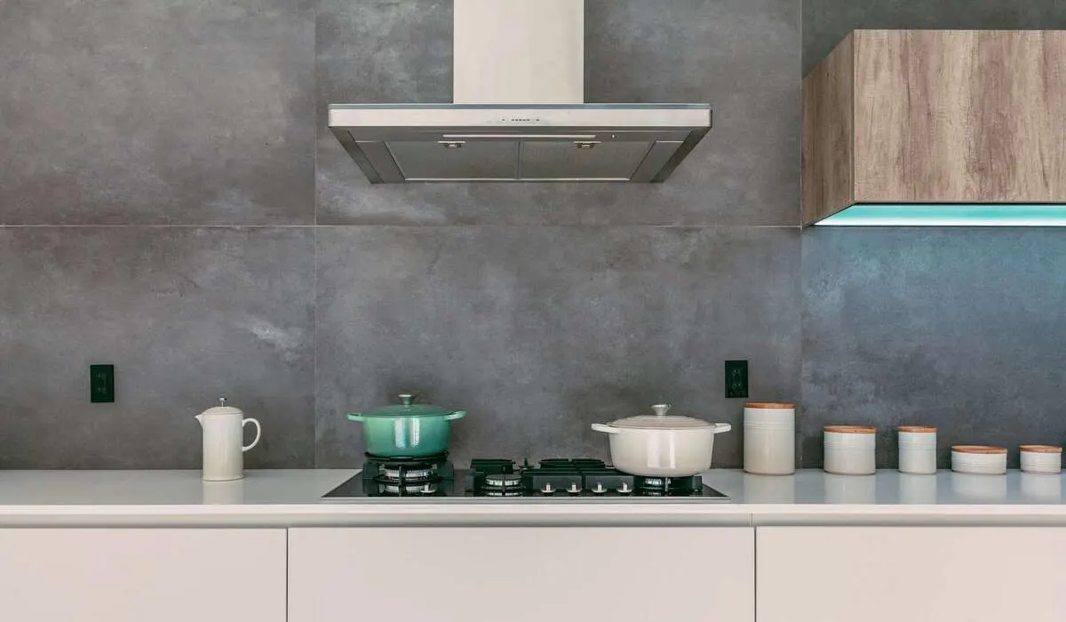 Cocina con ollas sobre hornilla, tetera blanca, recipientes y campana extractora bajo un armario de madera sobre una pared gris.