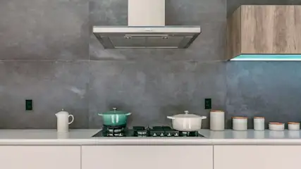 Cocina con ollas sobre hornilla, tetera blanca, recipientes y campana extractora bajo un armario de madera sobre una pared gris.