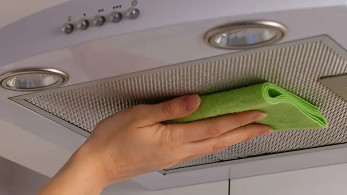 Una mano limpia el filtro metálico de una campana extractora blanca con un paño verde. La campana tiene luces y botones.