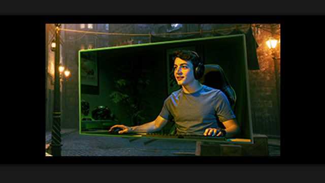 Un hombre con audífonos frente a un monitor con borde verde, usa teclado y ratón. Farolas iluminan un fondo urbano nocturno.