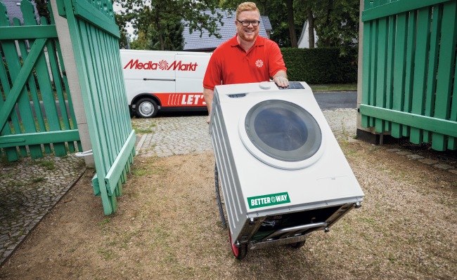 Un hombre con camisa roja mueve una lavadora blanca con un carro. Una furgoneta blanca y una valla verde están al fondo.
