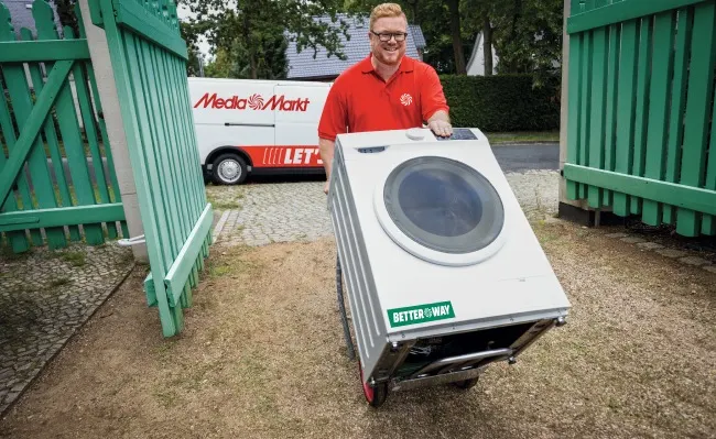 Un hombre con camisa roja mueve una lavadora blanca con un carro. Una furgoneta blanca y una valla verde están al fondo.