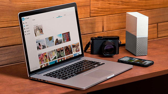 Un portátil abierto muestra fotos. Junto hay una cámara negra, un disco duro blanco y gris, y un teléfono móvil sobre una mesa de madera.