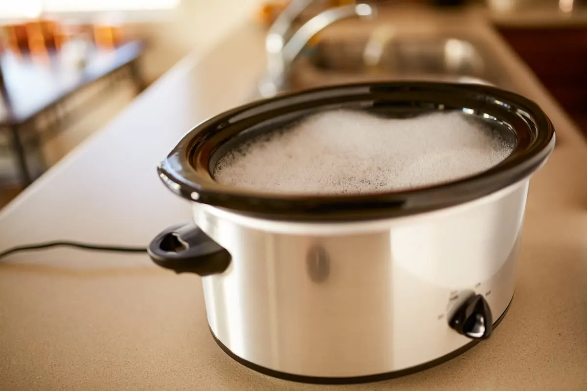 Olla de cocción lenta plateada sobre encimera de cocina con vapor visible bajo la tapa.