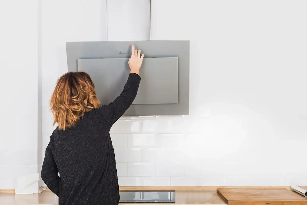 Una persona activa una campana extractora gris en una cocina blanca con azulejos y encimera de madera.