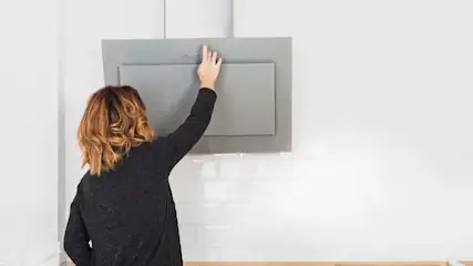 Una persona activa una campana extractora gris en una cocina blanca con azulejos y encimera de madera.