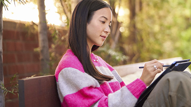 Una persona sentada en un banco usa una tableta con un lápiz en un entorno exterior con árboles y follaje.
