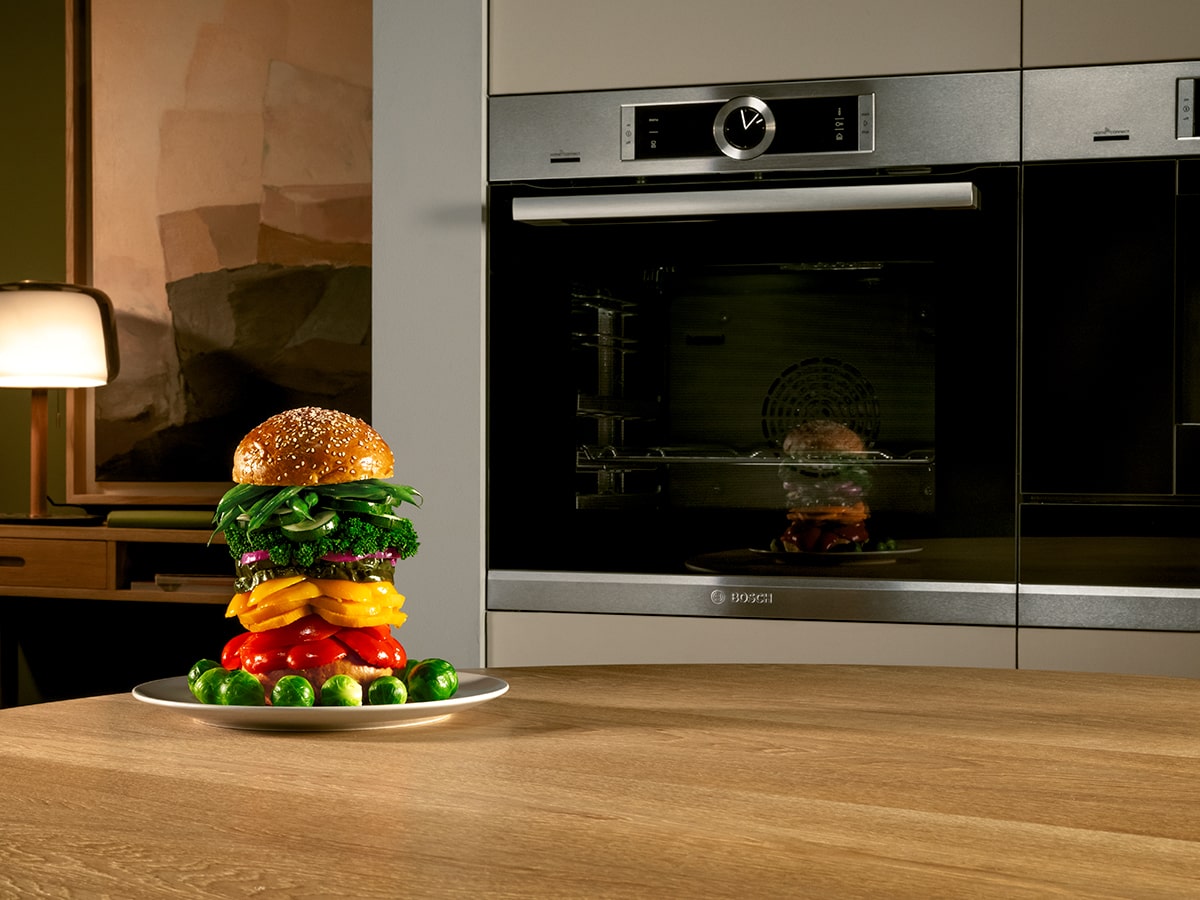 En una cocina, un plato con una hamburguesa vegetal alta y coles de Bruselas está sobre una mesa de madera frente a un horno moderno.