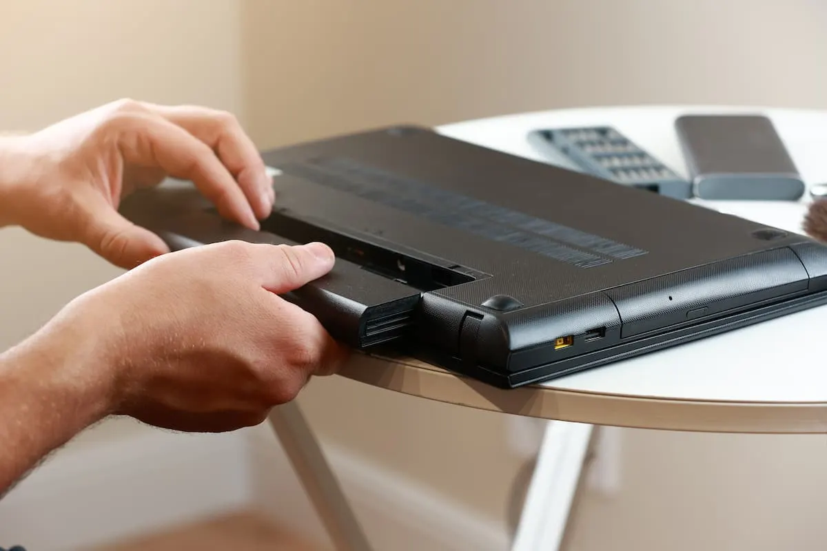 Manos de una persona extrayendo la batería de la parte inferior de un laptop negro colocado boca abajo sobre una mesa blanca.