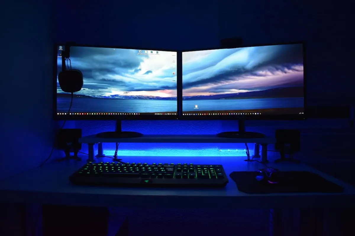 Escritorio gamer con dos monitores encendidos mostrando un paisaje, teclado retroiluminado y luz LED azul en un entorno oscuro.
