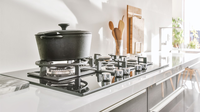Olla negra con tapa en estufa de gas. Utensilios de cocina de madera y tabla de cortar al fondo, junto a ventana luminosa.