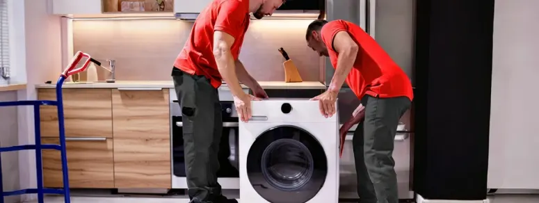 Dos técnicos con camiseta roja instalando una lavadora en una cocina