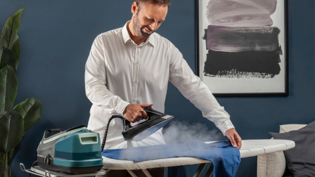 Mano sujetando plancha vertical negra y gris que emite vapor sobre tela gris. Persona vestida con camisa azul y vaqueros.
