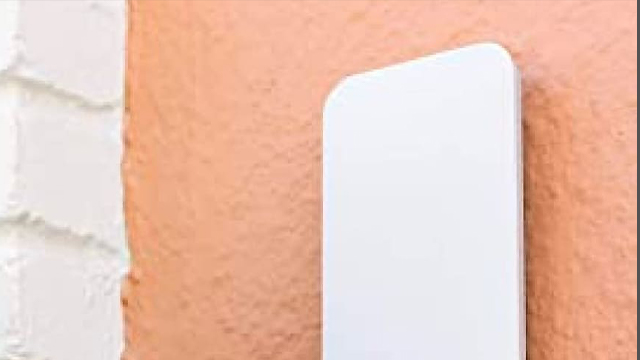 Un objeto blanco rectangular se apoya contra una pared de textura rugosa y color melocotón, junto a una pared blanca de ladrillo.