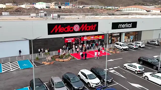 Tienda Media Markt con gente entrando por alfombra roja y letreros rojos. Aparcamiento lleno de coches y edificios al fondo.