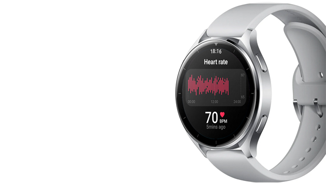 Reloj plateado con correa gris claro. La pantalla muestra datos de ritmo cardíaco con un gráfico rojo y el número 70 en blanco.