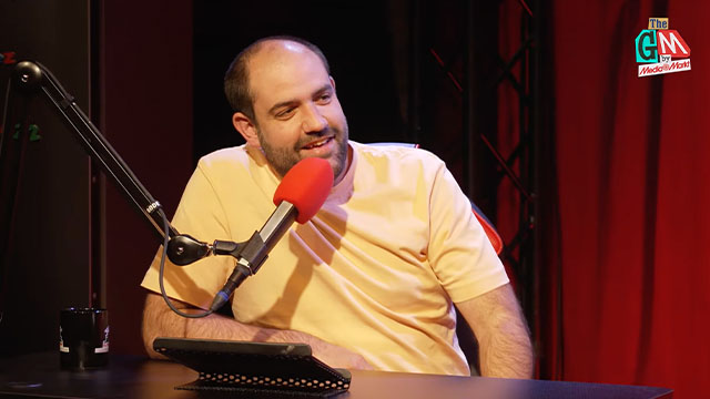 Un hombre en una mesa con un micrófono rojo, un teléfono y un vaso. Fondo de cortinas rojas y un logotipo.