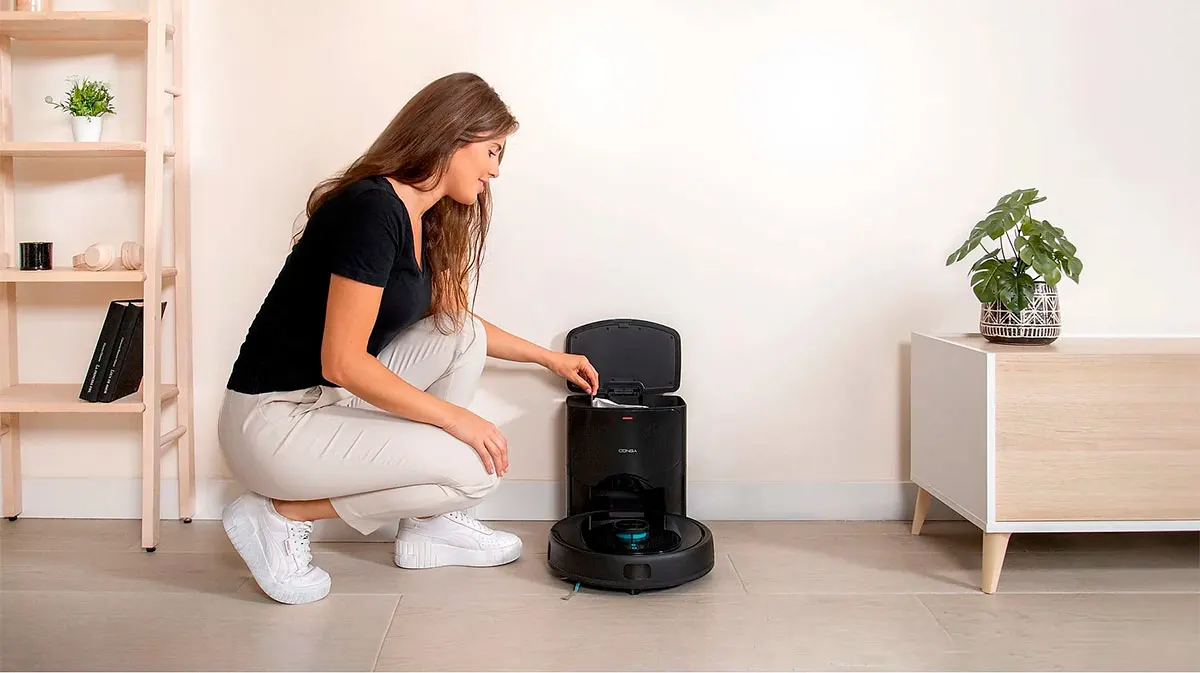 Mujer agachada junto a robot aspirador negro con base de carga abierta. Estantería de madera y planta decorativa al fondo.