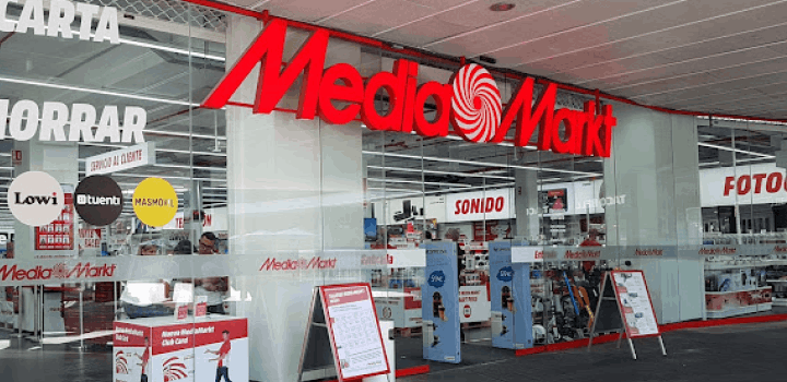 Entrada de tienda con letrero rojo "Media Markt", escaparate con ofertas, y carteles promocionales. Interior con secciones de sonido y fotografía.